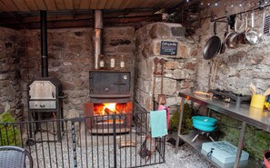 A rustic kitchen with a stone wall, a wood-burning stove, and cooking utensils hanging on the wall. - Railway Ramblers' Rest (Yelverton)