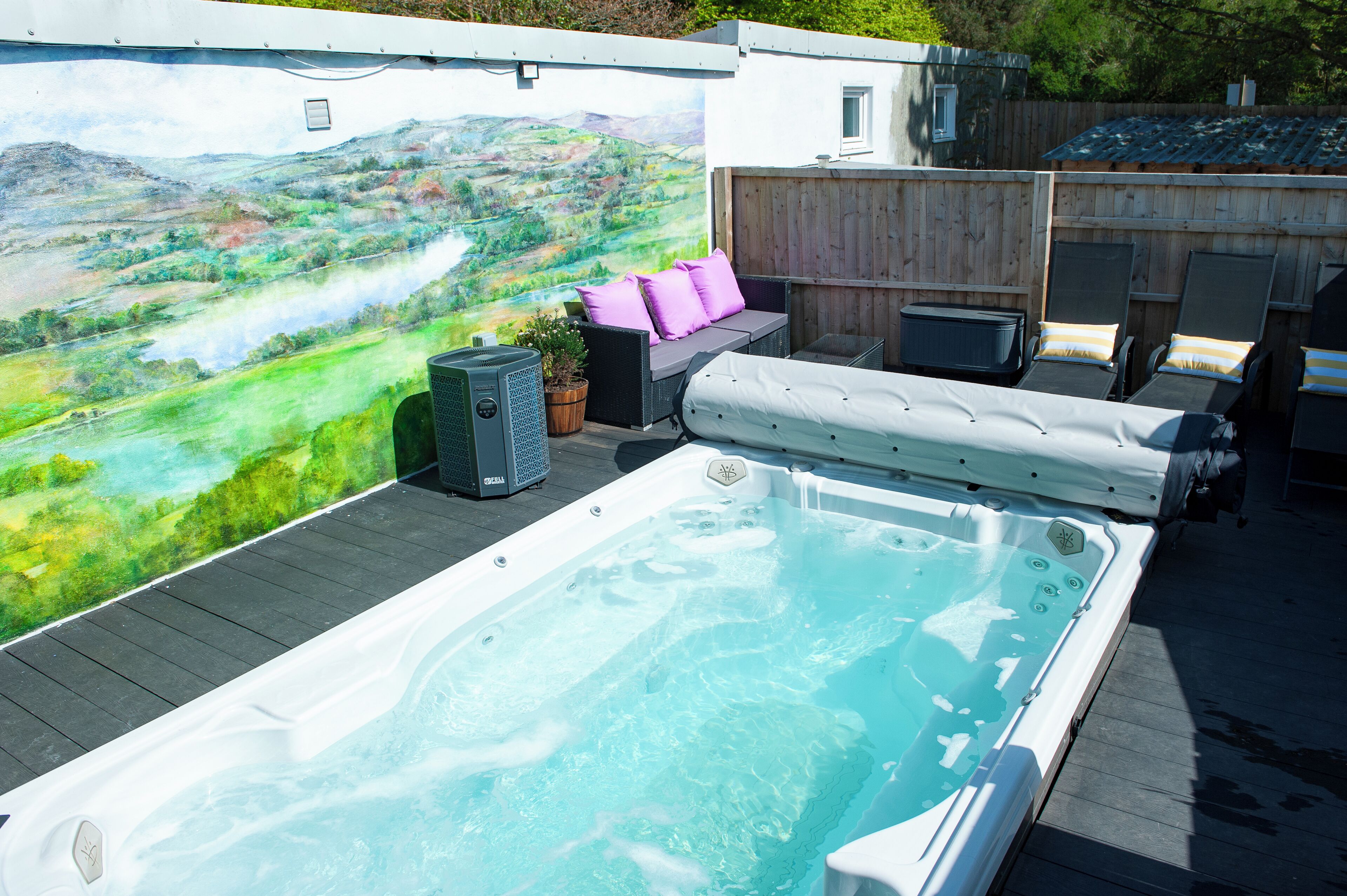 A hot tub with a wooden deck, a mural of a landscape, and outdoor seating with cushions.
