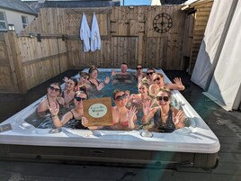 Un grupo de personas en una bañera de hidromasaje, con una valla de madera y un reloj al fondo.