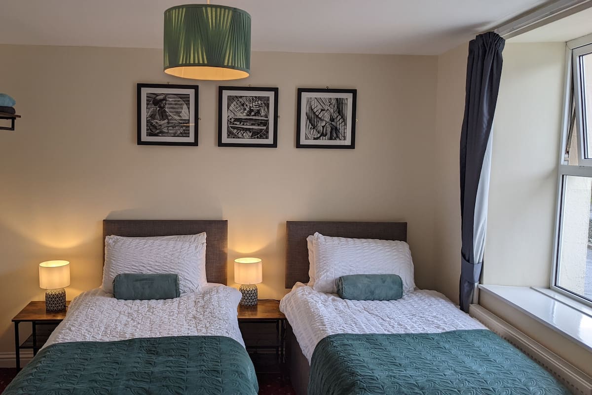 A hotel room with two beds, a window with curtains, a green lamp, and three framed pictures on the wall.
