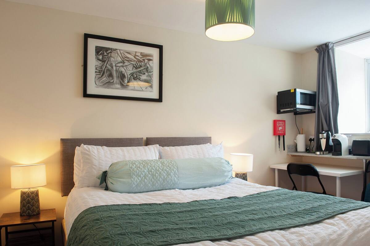 A hotel room with a bed, a desk, a chair, a lamp, a microwave, and a fire blanket.
