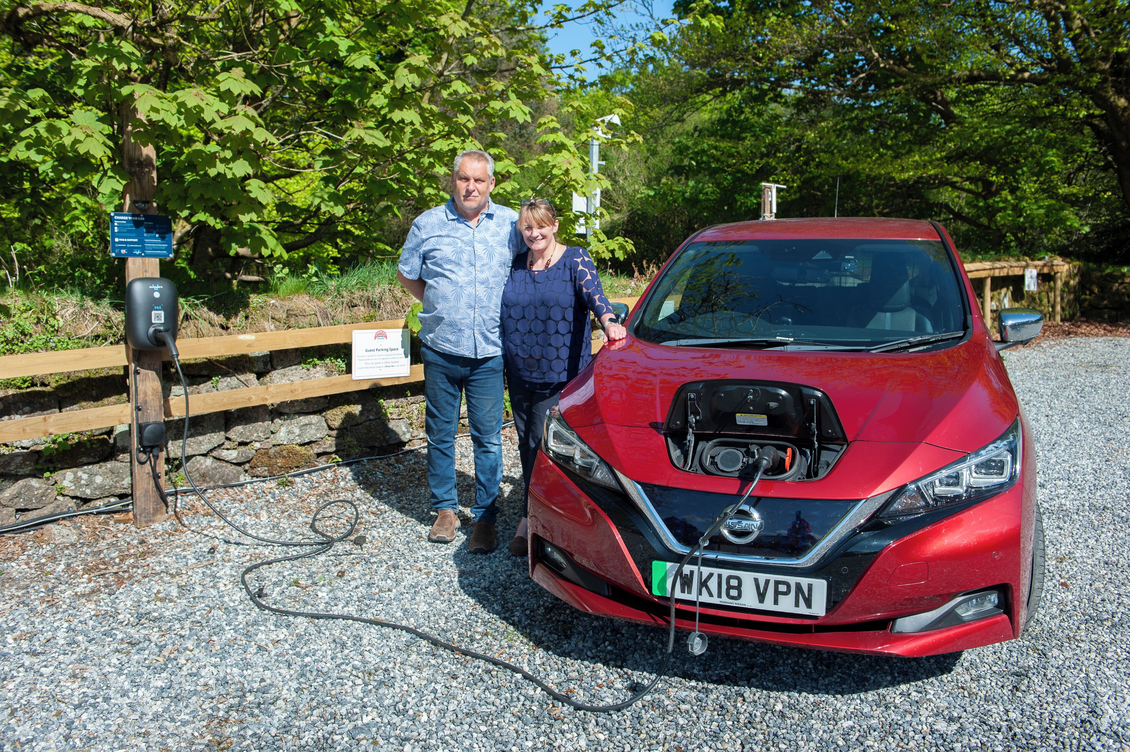 A red Nissan with a charging cable connected to a charging station, a wooden fence, and trees in the background.