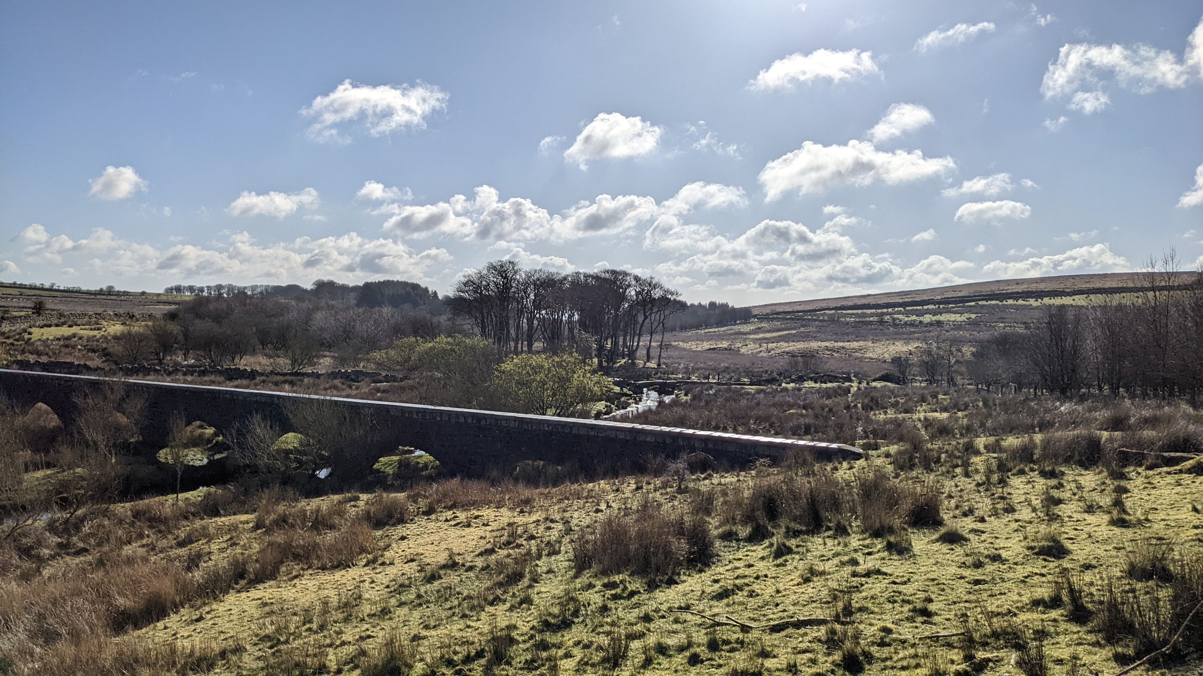 A rural landscape with a stone bridge, fields, and trees under a clear sky.