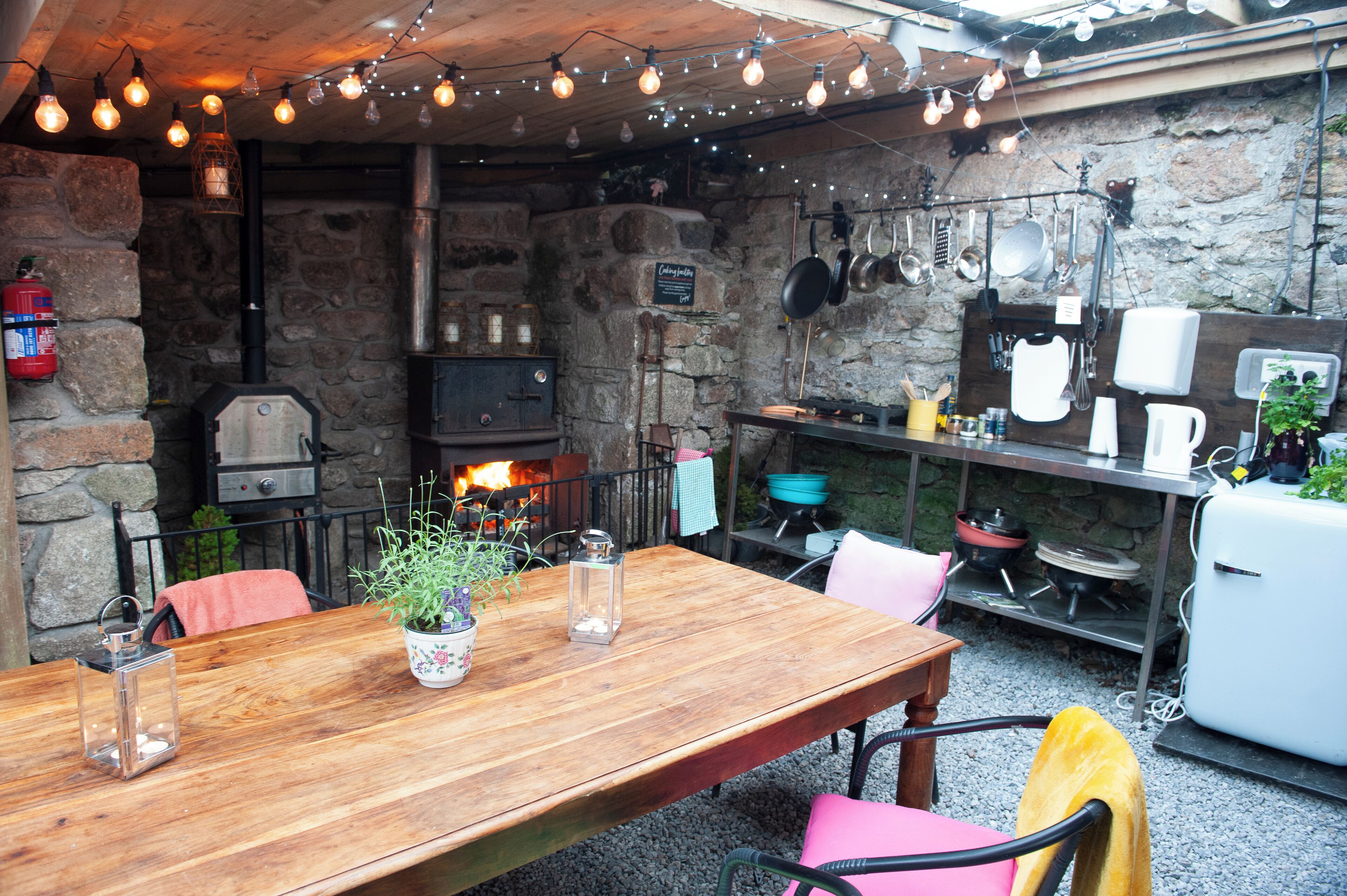 A rustic outdoor kitchen with a stone wall, wooden table, and various cooking utensils.