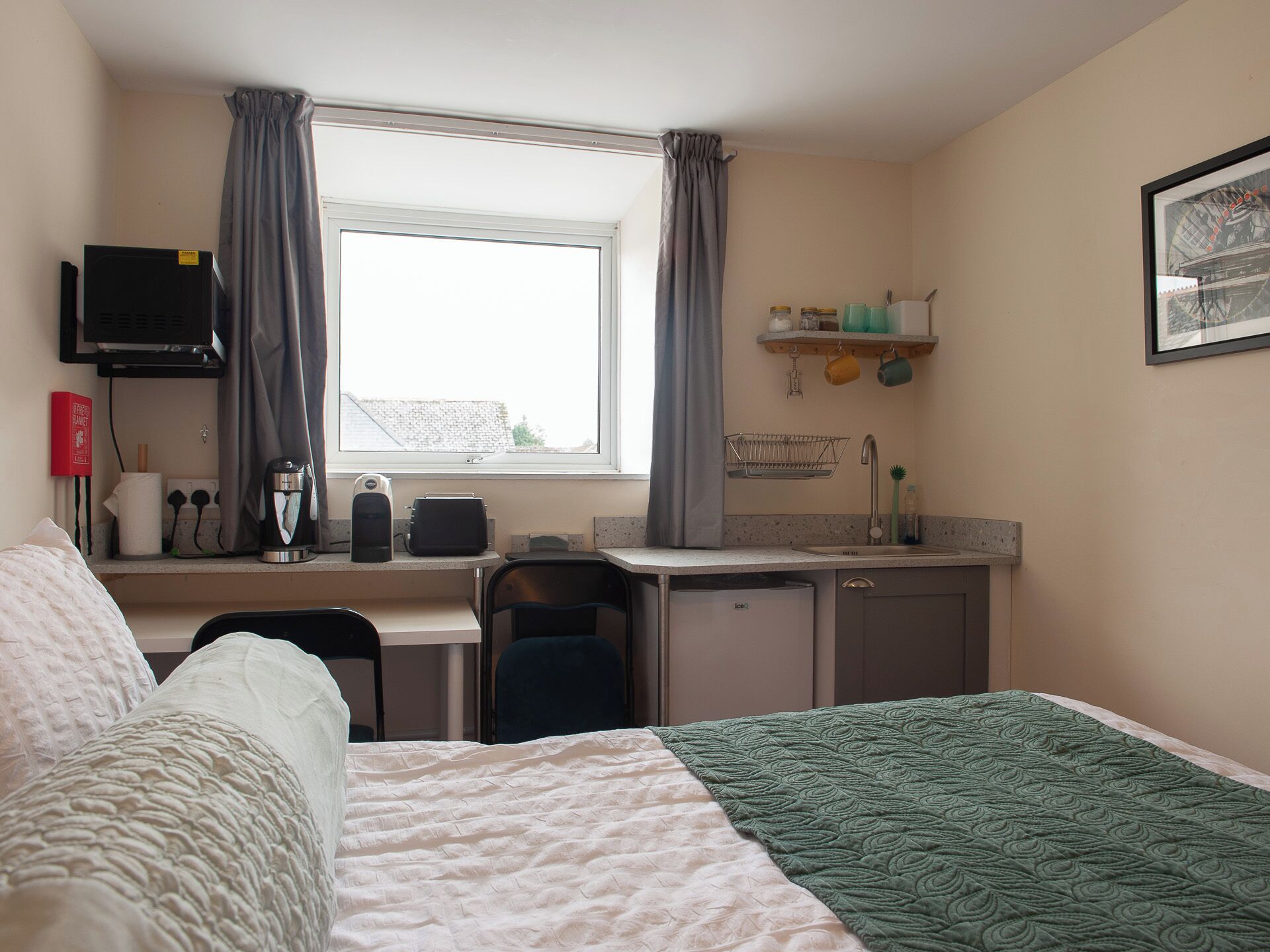 Habitación de hotel con cama, escritorio con computadora, silla, ventana con cortinas, lavamanos y estante con utensilios de cocina.