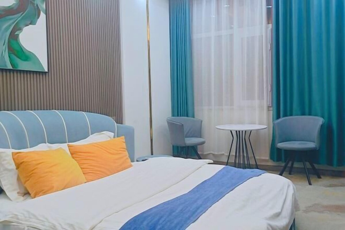 A hotel room with a bed, a blue upholstered headboard, two chairs, a small round table, and a large window with curtains.