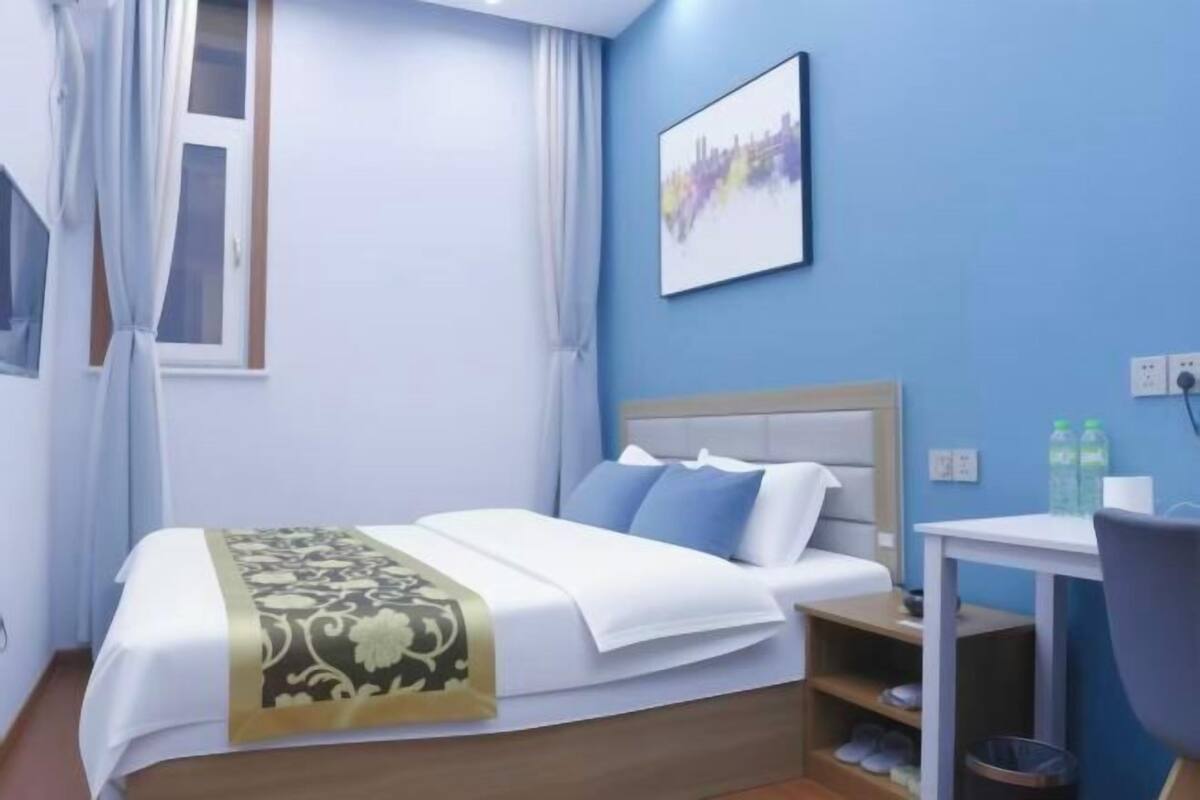 Ett hotellrum med en säng, ett skrivbord, en stol, ett fönster med gardiner, en tavla som hänger på väggen och ett litet bord med skor på.