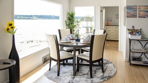 Un coin repas avec vue sur l’eau, une table ronde dressée pour quatre personnes et un vase rempli de tournesols placé sur une table d’appoint.