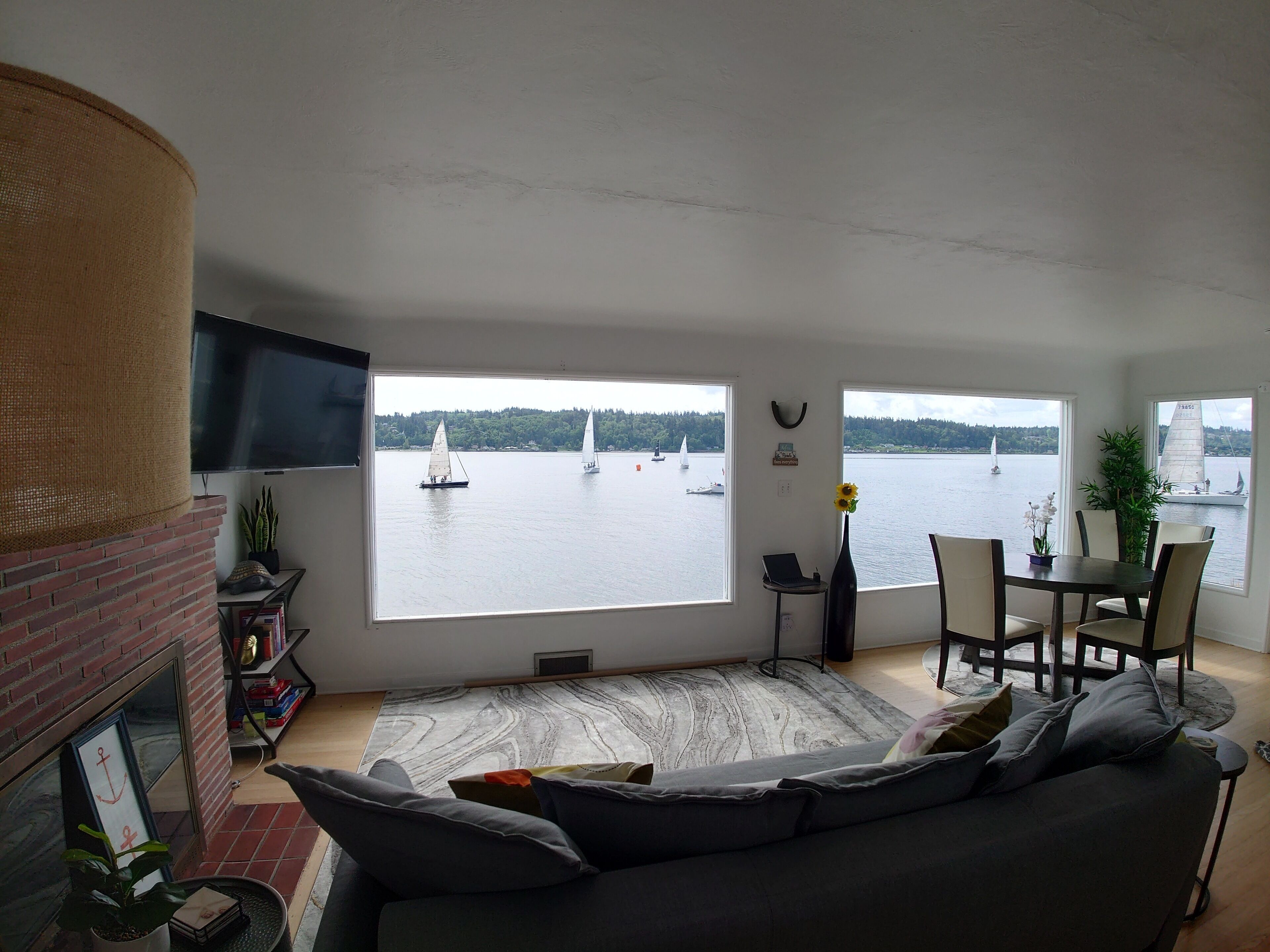 En stue med stort vindue der udover en sø med sejlbåde, en pejs, et fjernsyn og et spiseområde med stole og et bord.