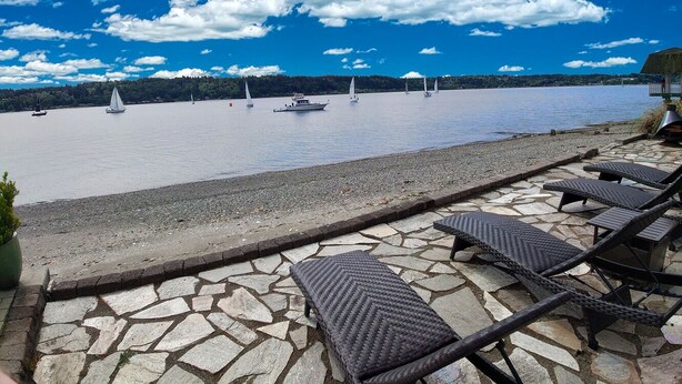 Une vue sur le front de mer, avec des chaises longues et une terrasse en pierre donnant sur une étendue d’eau calme où voguent des voiliers.