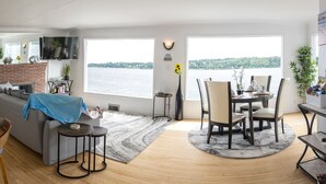 Un salon moderne avec une cheminée, un coin repas offrant une vue sur l’eau et une cuisine.
