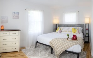 Une chambre à coucher comprenant un lit, des tables de chevet, une commode et une fenêtre avec des rideaux légers.