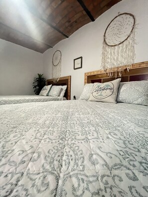 Ein Zimmer mit zwei Betten, jeweils mit einem hölzernen Kopfteil und einer gemusterten Bettdecke. An der Wand hängen zwei Traumfänger, darüber ein kleines gerahmtes Bild.