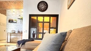 Ein gemütliches Wohnzimmer mit einem Holztisch, einem beigefarbenen Sofa und einem blauen Kissen sowie einer Uhr an der Wand.