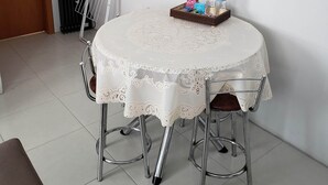 Una mesa de comedor con mantel de encaje, rodeada de sillas de metal, en una habitación con paredes blancas y piso de baldosas.