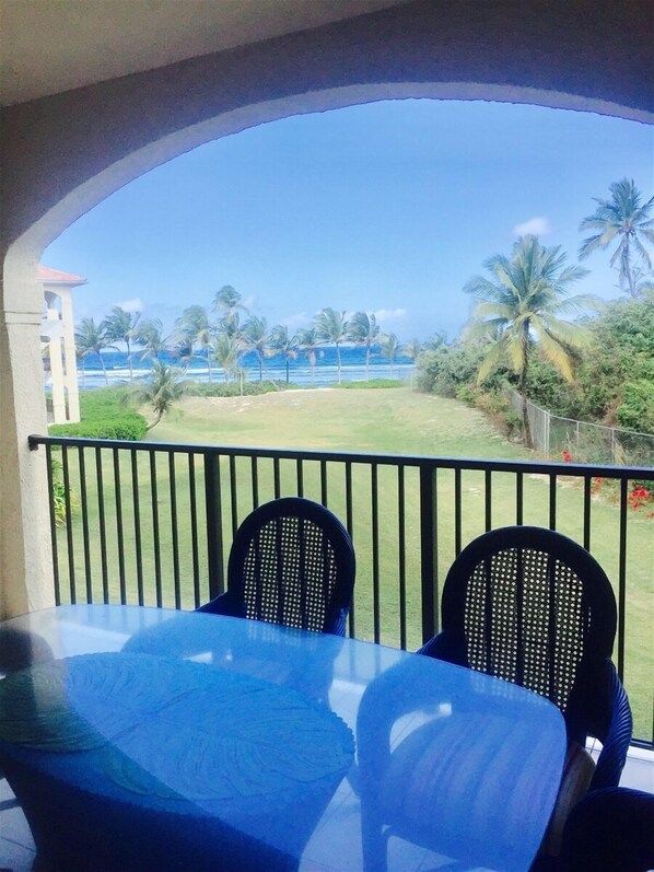 Varanda com duas cadeiras e uma mesa, com vista para uma praia com palmeiras e céu claro.
