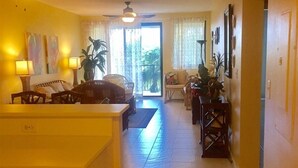 Un'ampia zona giorno con cucina, tavolo da pranzo e sedie. È presente un'ampia finestra con tende, una ventola a soffitto e una porta che conduce in un'altra stanza.