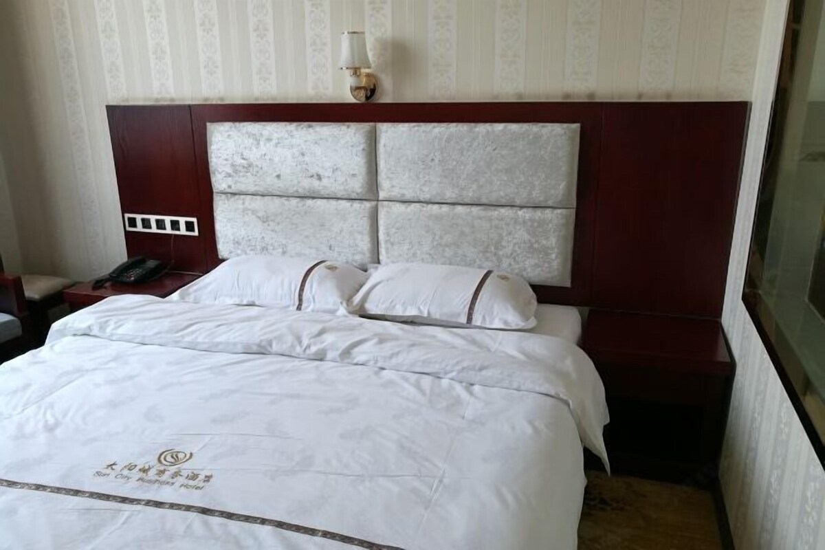 A hotel room with a large bed, a headboard with a textured design, a bedside table with a lamp, and a phone on the table.