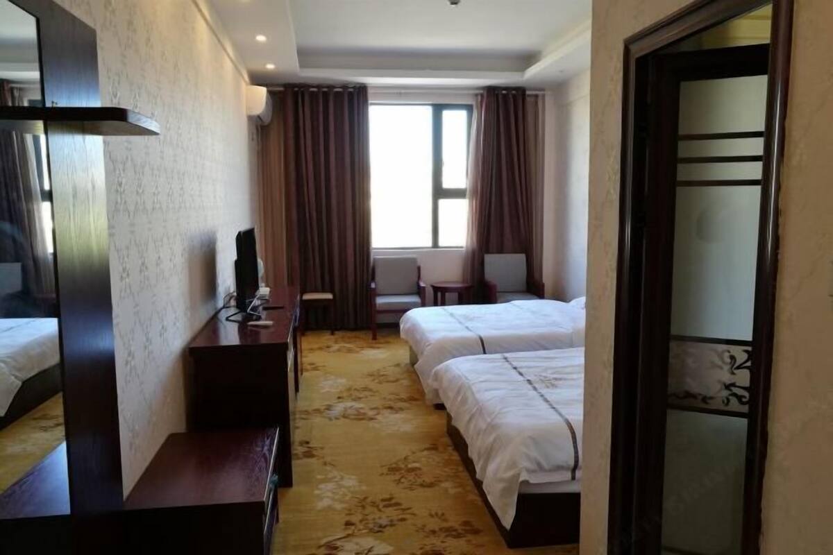 A hotel room with two beds, a desk, a chair, a TV, and a large window with curtains.