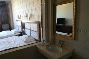 A hotel room with a bed, a bathroom with a sink, a mirror, and a television in the corner. - Sun City Business Hotel (Yongren)