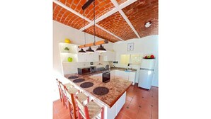 Una cocina con una isla central, techo de ladrillo rojo, armarios blancos y suelo de baldosas.