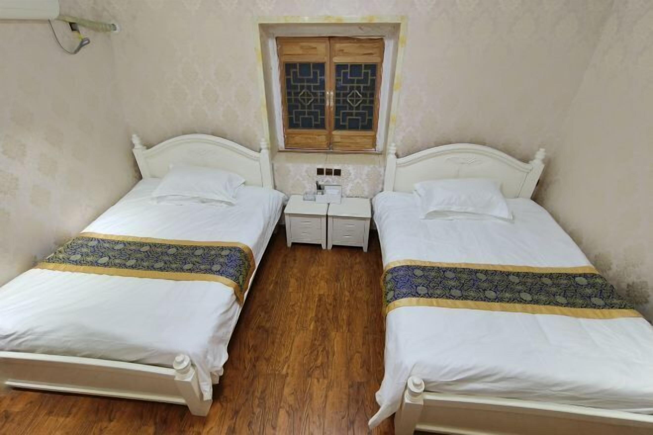 Ein Zimmer mit zwei Einzelbetten, Holzboden, einem kleinen Nachttisch mit Lampe und einem Fenster mit Dekorglas.