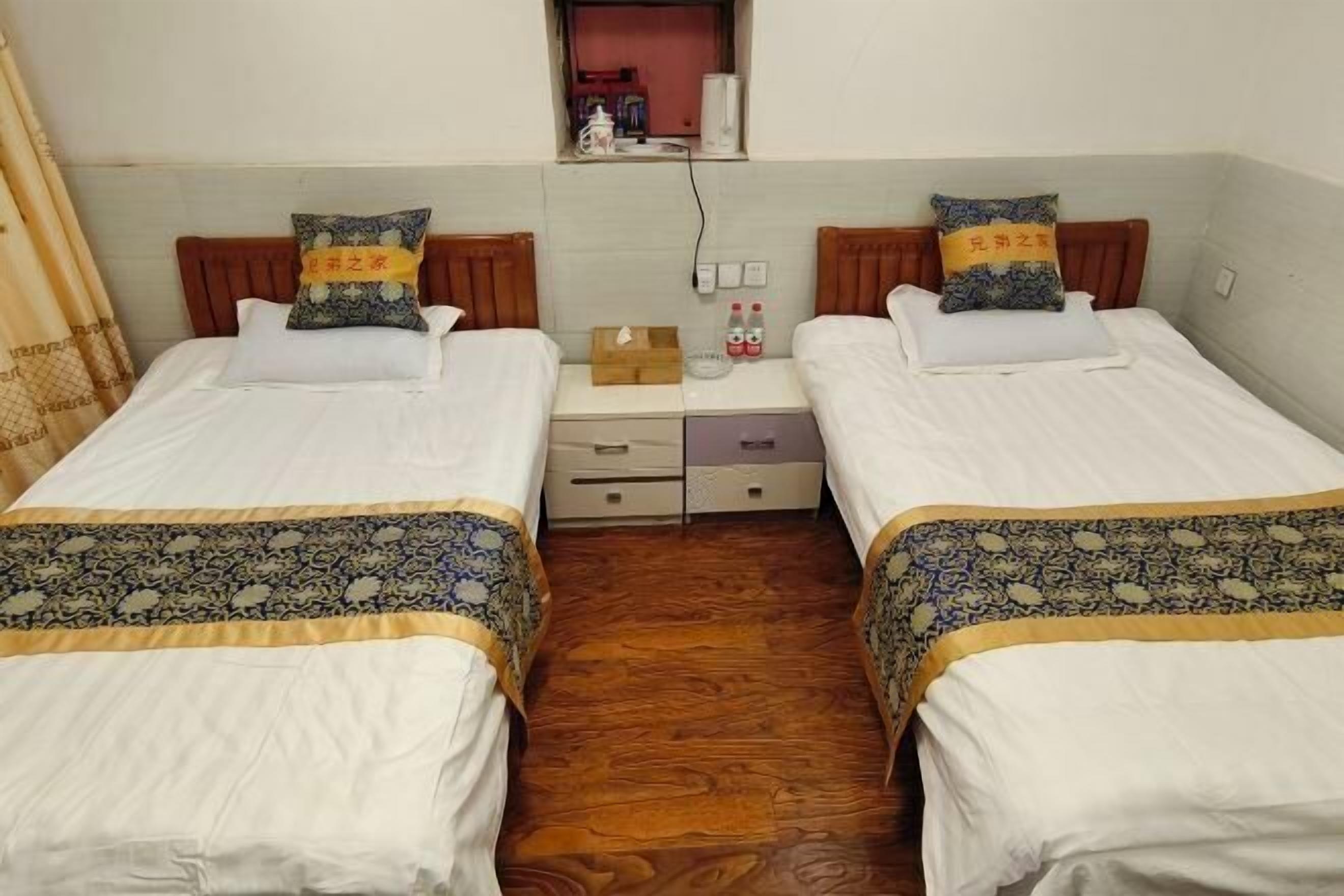 A room with two beds, each with a patterned bedspread and pillows with text. There is a small table with a box, bottles, and a lamp between the beds.