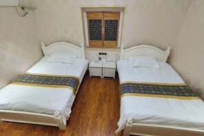Une petite chambre propre, avec deux lits simples, une fenêtre en bois ornée de vitraux et une table de chevet blanche placée entre les lits.
