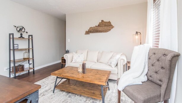 A living room with a wooden coffee table, a white sofa, a beige armchair, and a metal shelf unit with decorative items.