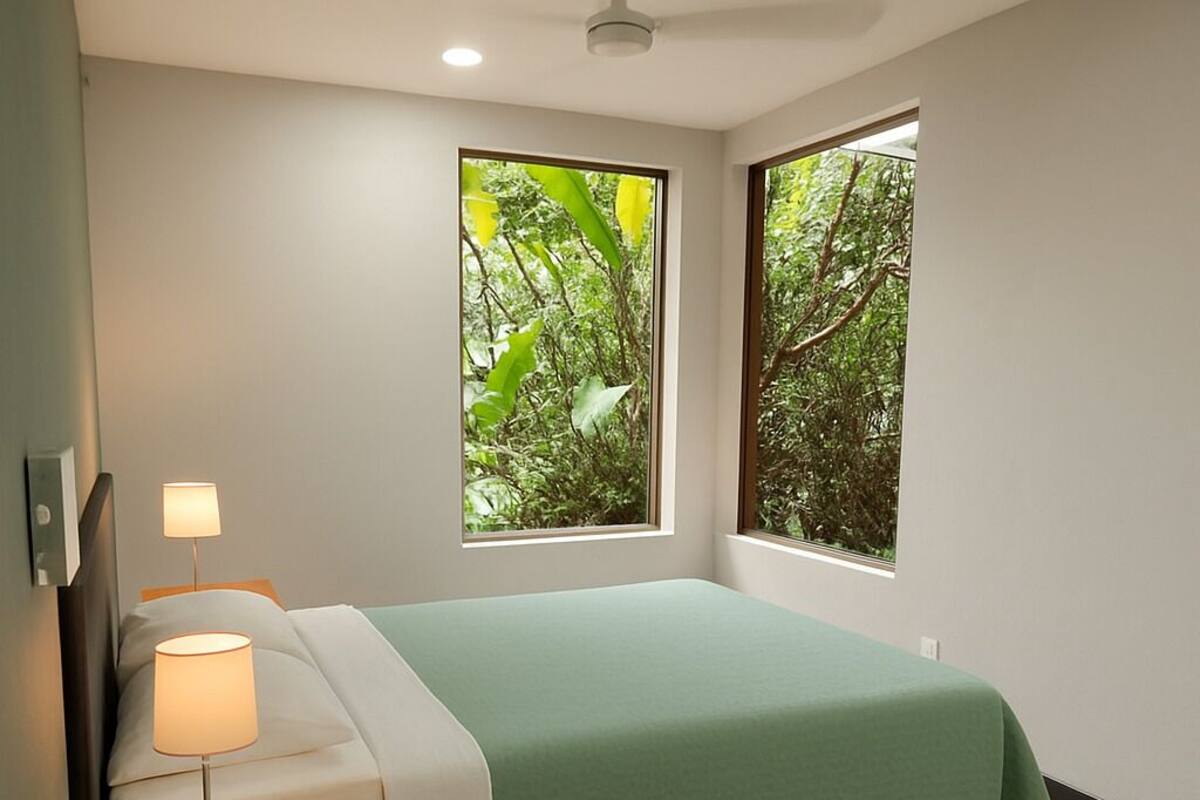 Une chambre à coucher comprenant un lit, des lampes de chevet, une table de chevet, une fenêtre donnant sur un espace verdoyant et un luminaire au plafond.