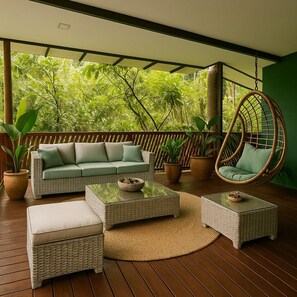 A covered outdoor seating area with wicker furniture, including a sofa, two ottomans, and a hanging chair, surrounded by potted plants and a view of lush greenery. - Green Jungle House (Cahuita)