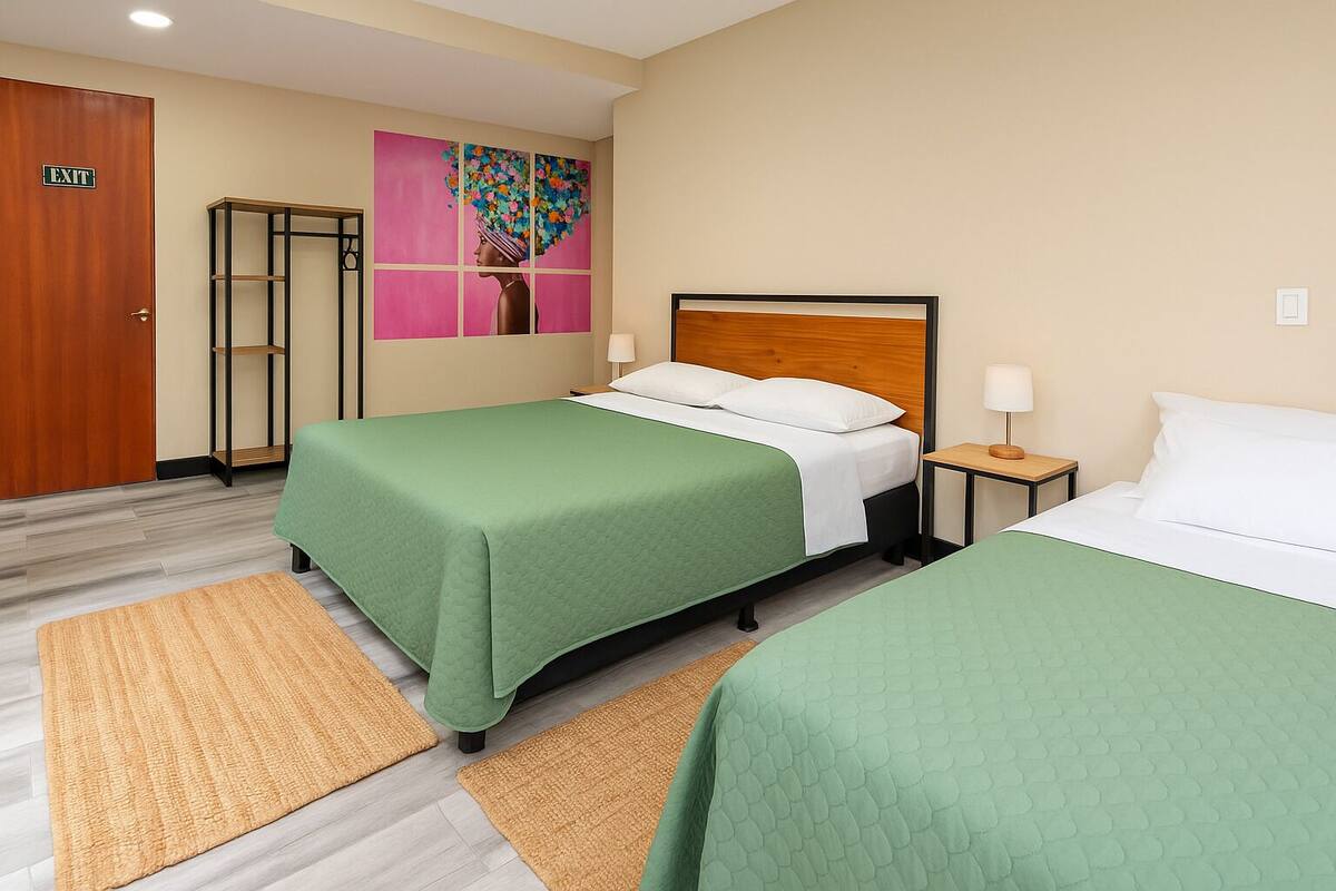 Une chambre d’hôtel avec deux lits, une couvre-lit verte, une tête de lit en bois, une table de chevet avec une lampe, un tableau au mur et une porte de sortie.