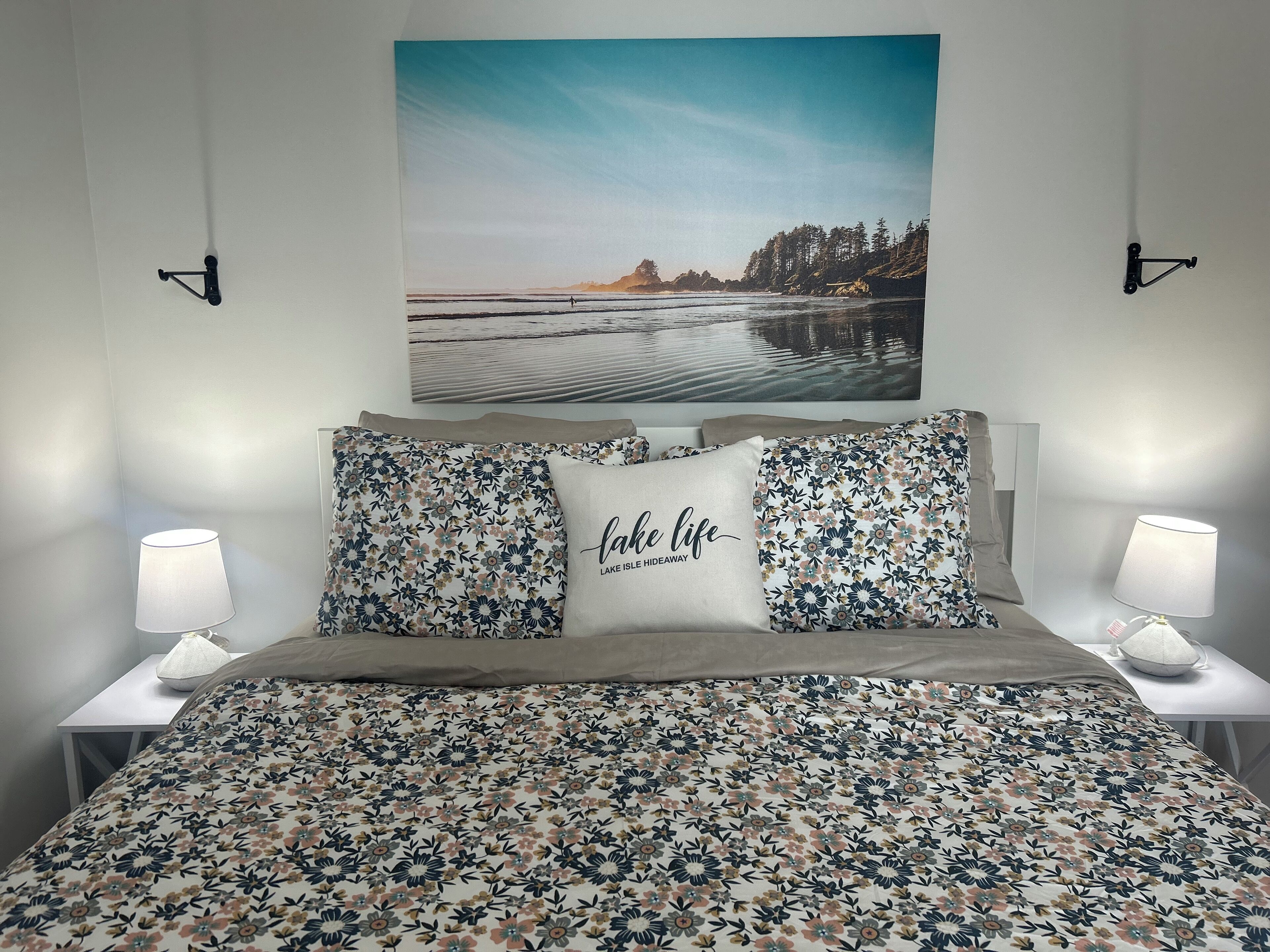 A bedroom with a floral bedspread, two matching pillows, a framed landscape painting, and bedside lamps.