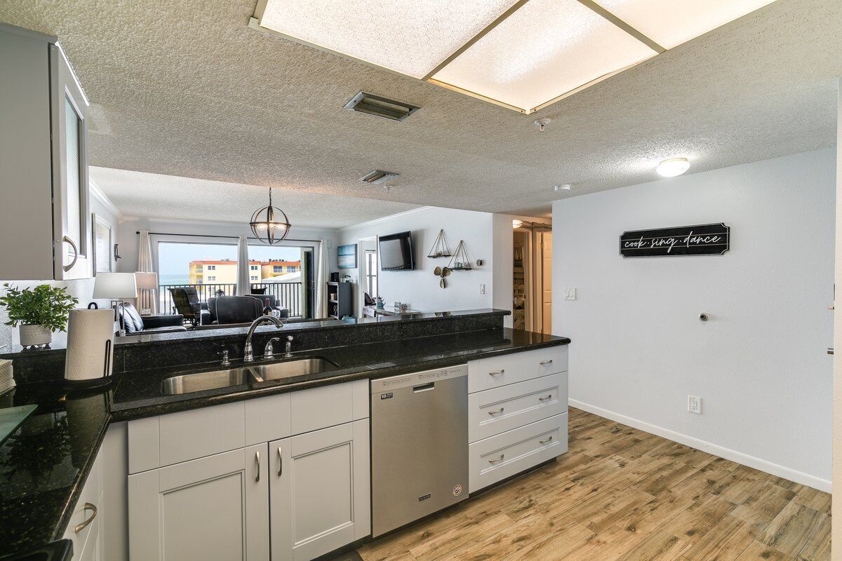 A modern kitchen with a black countertop, white cabinets, and a dishwasher. There is a large window providing a view of buildings outside.