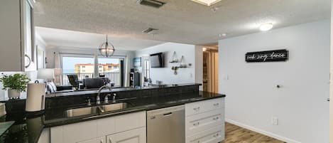 A modern kitchen with a black countertop, white cabinets, and a dishwasher. There is a large window providing a view of buildings outside.
