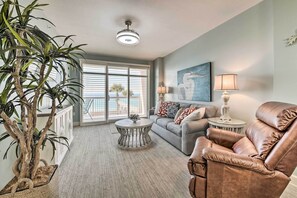 A living room with a brown leather chair, a gray sofa, a round coffee table, a floor lamp, and a large window with blinds. - Beachfront Condo w/Pool, Hot Tub & Beach Chairs Included (Panama City Beach)