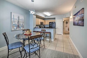 A modern kitchen with wooden cabinets, a black refrigerator, and a dining area with a glass table and blue patterned chairs.