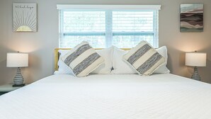 A neatly made bed with striped pillows, a bedside lamp, and a window with blinds. - Newly Renovated Home w/Outdoor Shower, Pool & Tiki Hut!  (Crystal River)