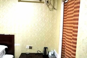 A hotel room with a bed, a small table with a kettle and cups, a power outlet, and a patterned curtain.