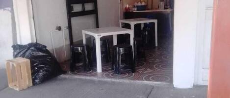 Un garaje con una mesa blanca y sillas negras, una bolsa de basura negra y un taburete de madera.