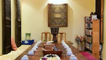 A long dining table with teapots and a fruit bowl, surrounded by chairs, in a room with traditional decor and a bookshelf.