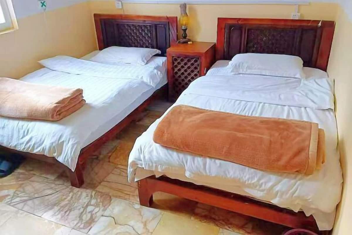 A room with two wooden beds, each with white bedding and a brown blanket. There is a small wooden nightstand with a lamp between the beds.