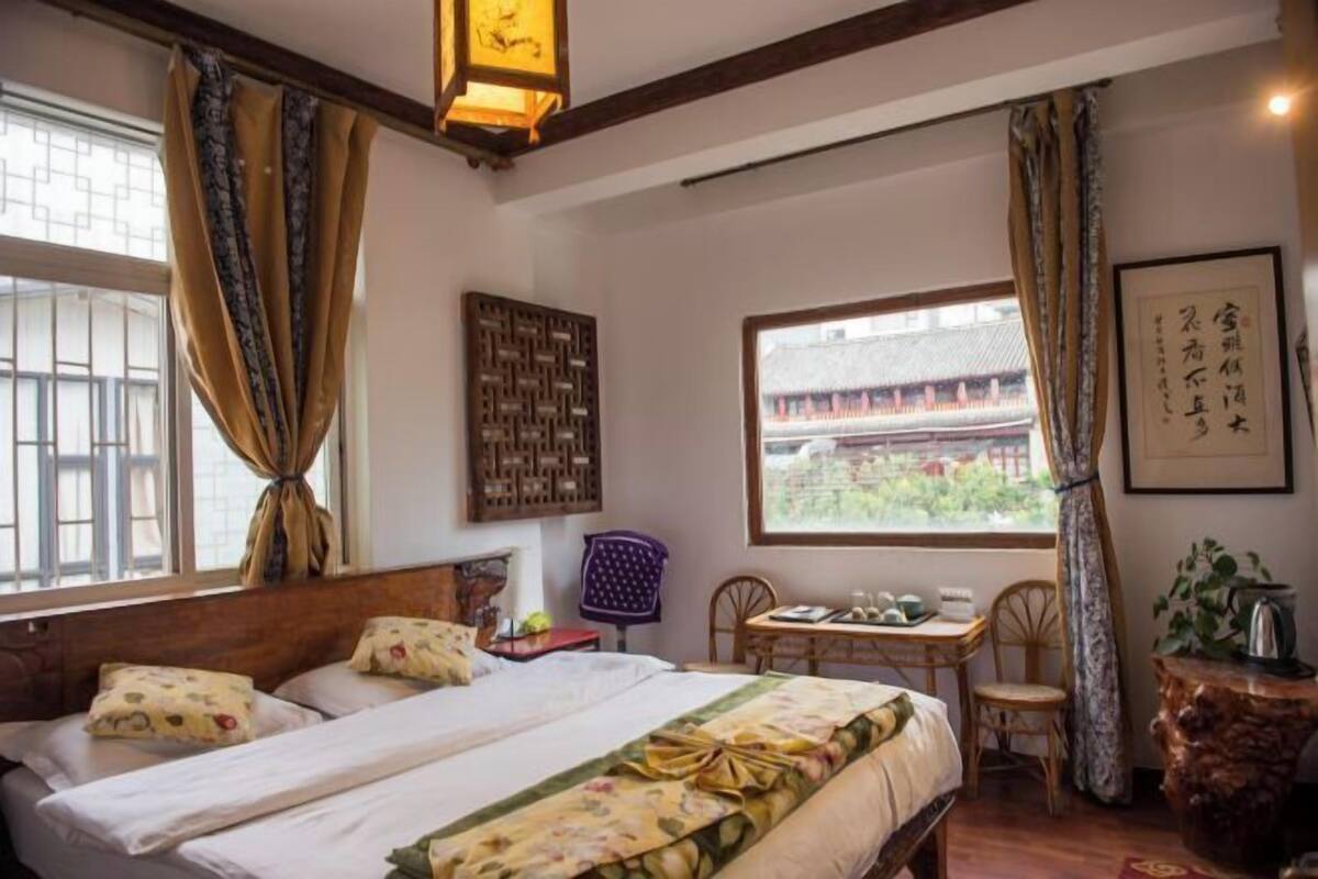 A traditional room with a bed, a small table with chairs, a window with a view of a traditional building, and a decorative wall piece.