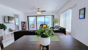 A modern living room with a dining table, sofa, and large windows offering a view of the outdoors. - Five O'clock Somewhere Condo (Northside)