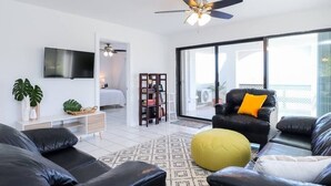 A modern living room with a black leather sofa, a yellow ottoman, and a black armchair with an orange pillow. A ceiling fan is visible, and there is a flat-screen TV mounted on the wall. - Five O'clock Somewhere Condo (Northside)