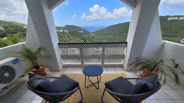 A balcony with two black chairs, a small round table, and a patterned rug. There are two potted plants and a white lattice partition. The balcony overlooks a green landscape with hills and a clear sky. - Five O'clock Somewhere Condo (Northside)