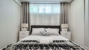 Una cama bien hecha con un edredón blanco y negro estampado, dos mesitas de noche con lámparas y una ventana con cortinas decoradas.