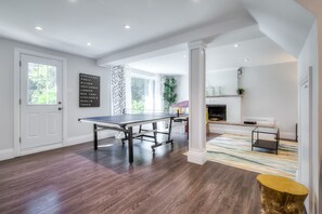 A modern living room with a ping pong table, a fireplace, and a view of greenery outside.