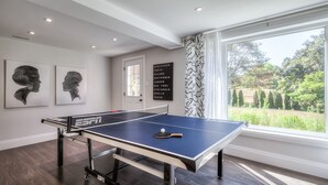 Una mesa de ping pong en una habitación con un ventanal grande, una puerta y dos retratos enmarcados en la pared.