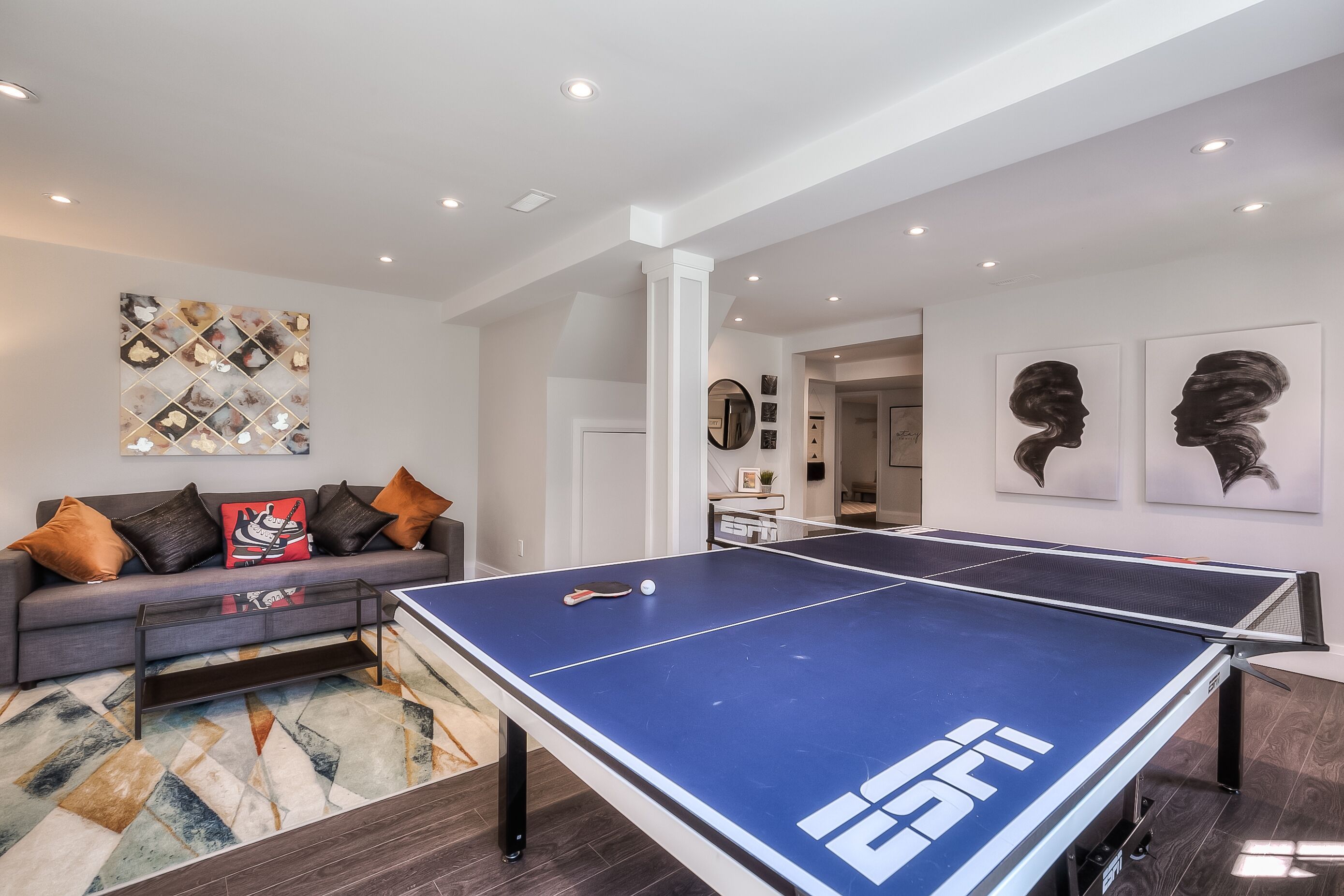 A modern living room with a ping pong table, a gray sofa with decorative pillows, and abstract wall art.